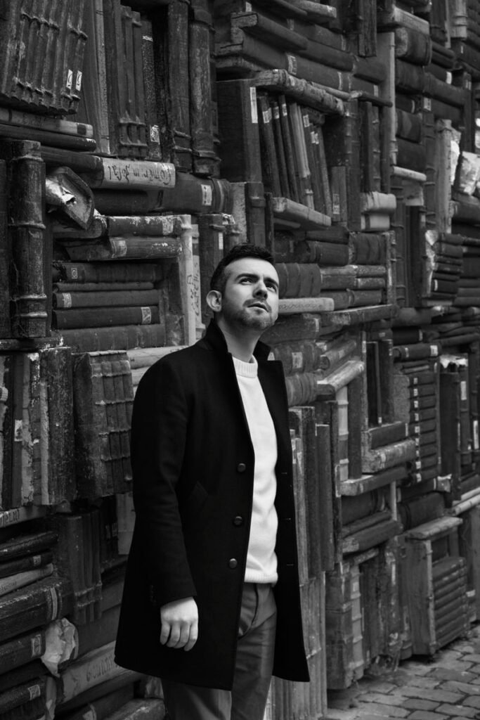 Marco Pennella, consulente di comunicazione, in cappotto scuro davanti a un muro di vecchi libri